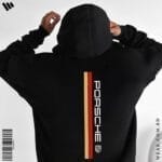 Back view of a man wearing a black oversized hoodie featuring a vertical red and gold stripe, the Porsche logo text, and the Porsche crest.