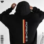 Back view of a man wearing a black oversized hoodie featuring a vertical red and gold stripe, the Porsche logo text, and the Porsche crest.