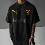 Front view of a man wearing a black oversized t-shirt with a gold Puma logo, a gold Scuderia Ferrari shield logo, and a tonal dark grey "Ferrari" text logo.