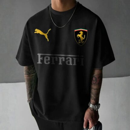 Front view of a man wearing a black oversized t-shirt with a gold Puma logo, a gold Scuderia Ferrari shield logo, and a tonal dark grey "Ferrari" text logo.